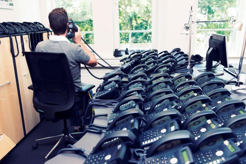 Man on phone surrounded by numerous telephones illustrating telecom cost management solutions.